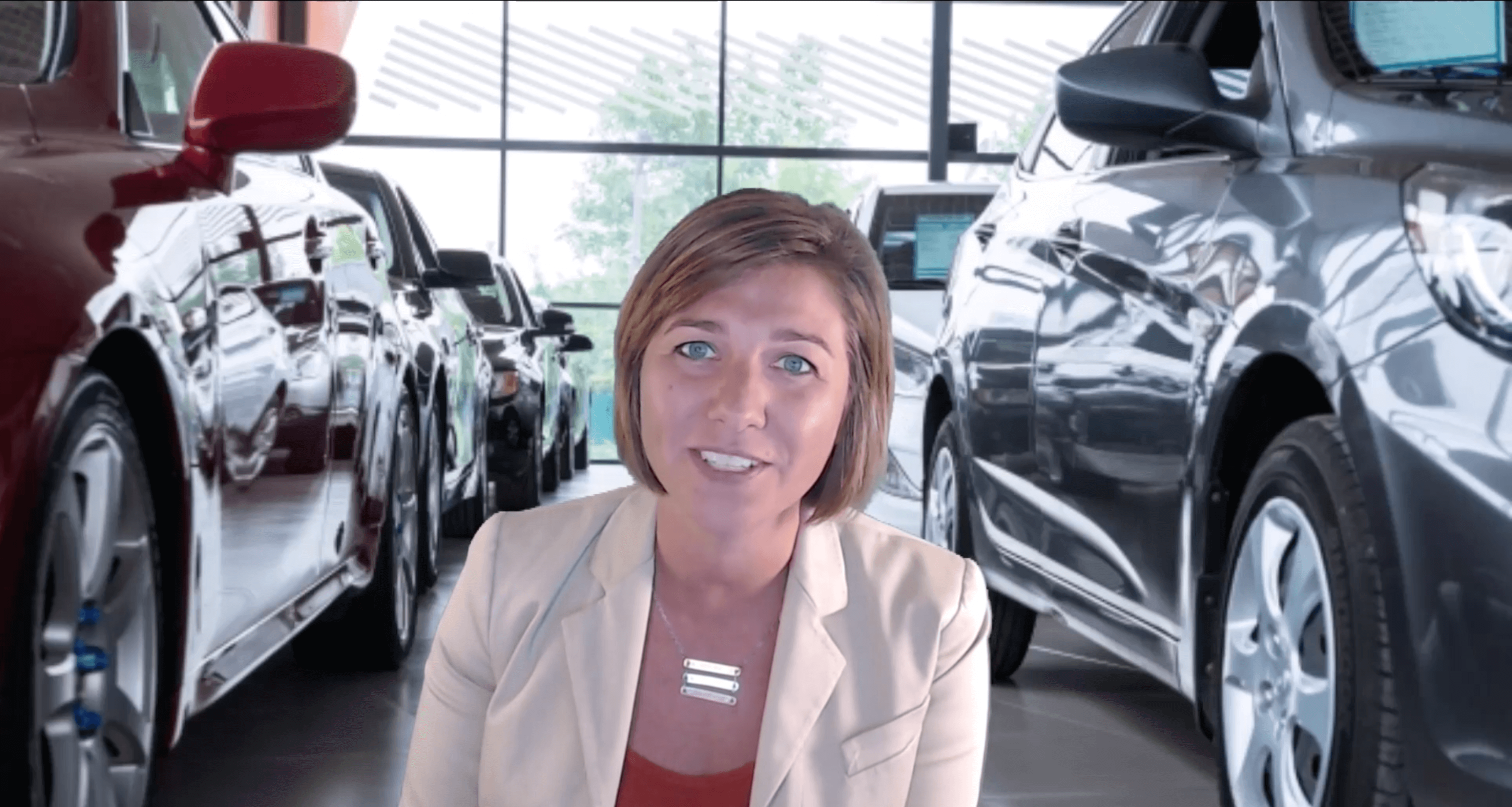 A person with a blurred face is standing in a car showroom, surrounded by various cars. The background shows large windows with natural light coming in.